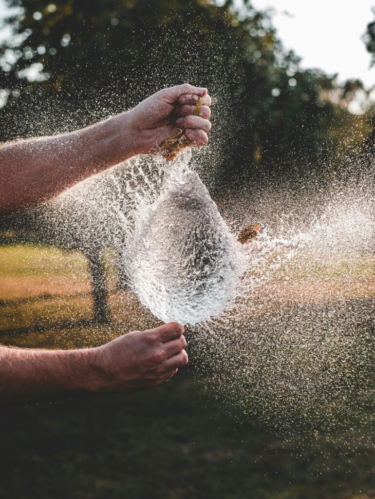 Two adult hands bursting a water balloon outdoors, captured mid-air, showing a dynamic splash.