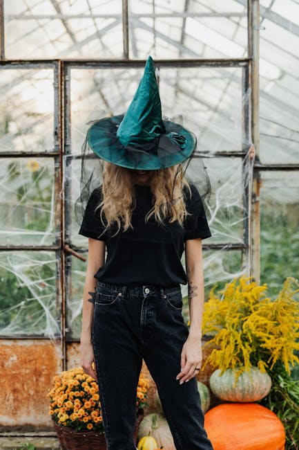 A woman in a witch costume stands in a greenhouse surrounded by pumpkins and flowers, perfect for Halloween themes.