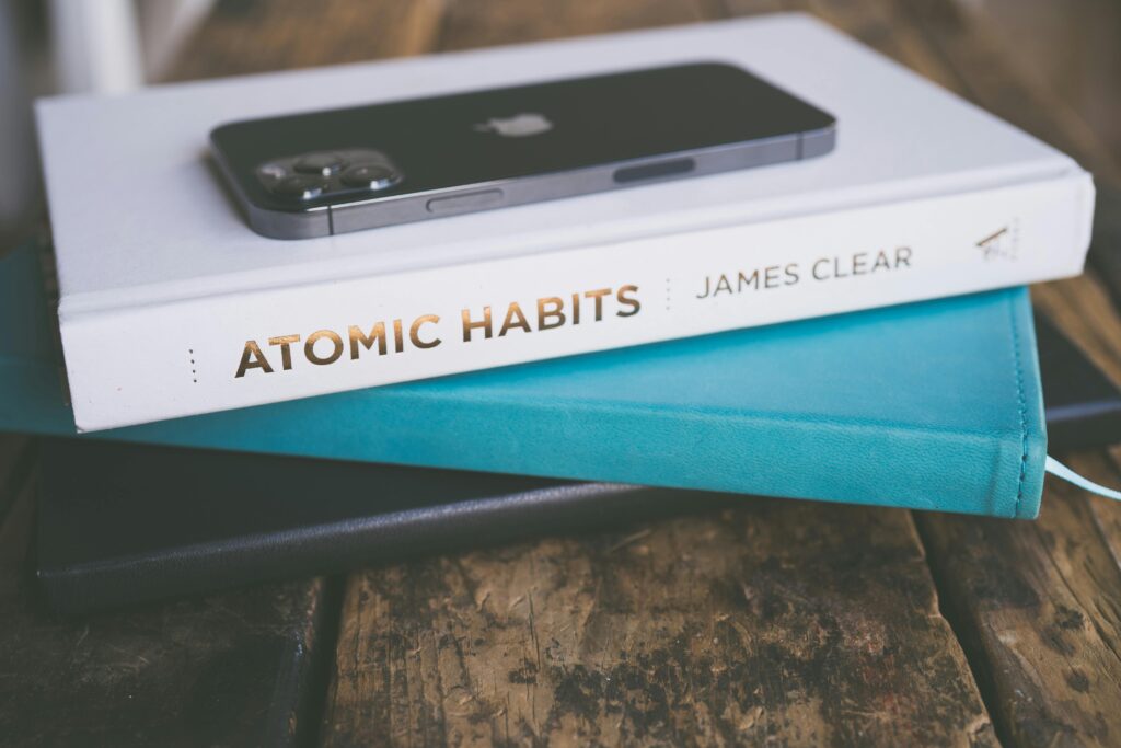 Close-up of a smartphone on a book stack, featuring 'Atomic Habits' by James Clear on a wooden surface.