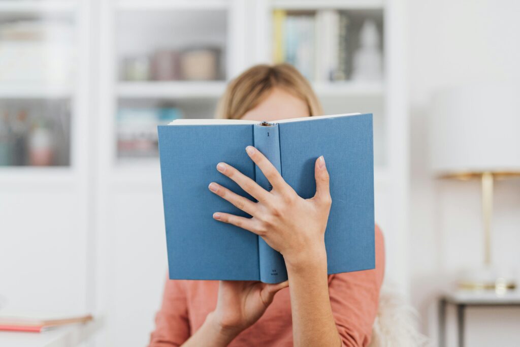 A woman holds and reads a blue book in a cozy indoor setting with a blurred background.